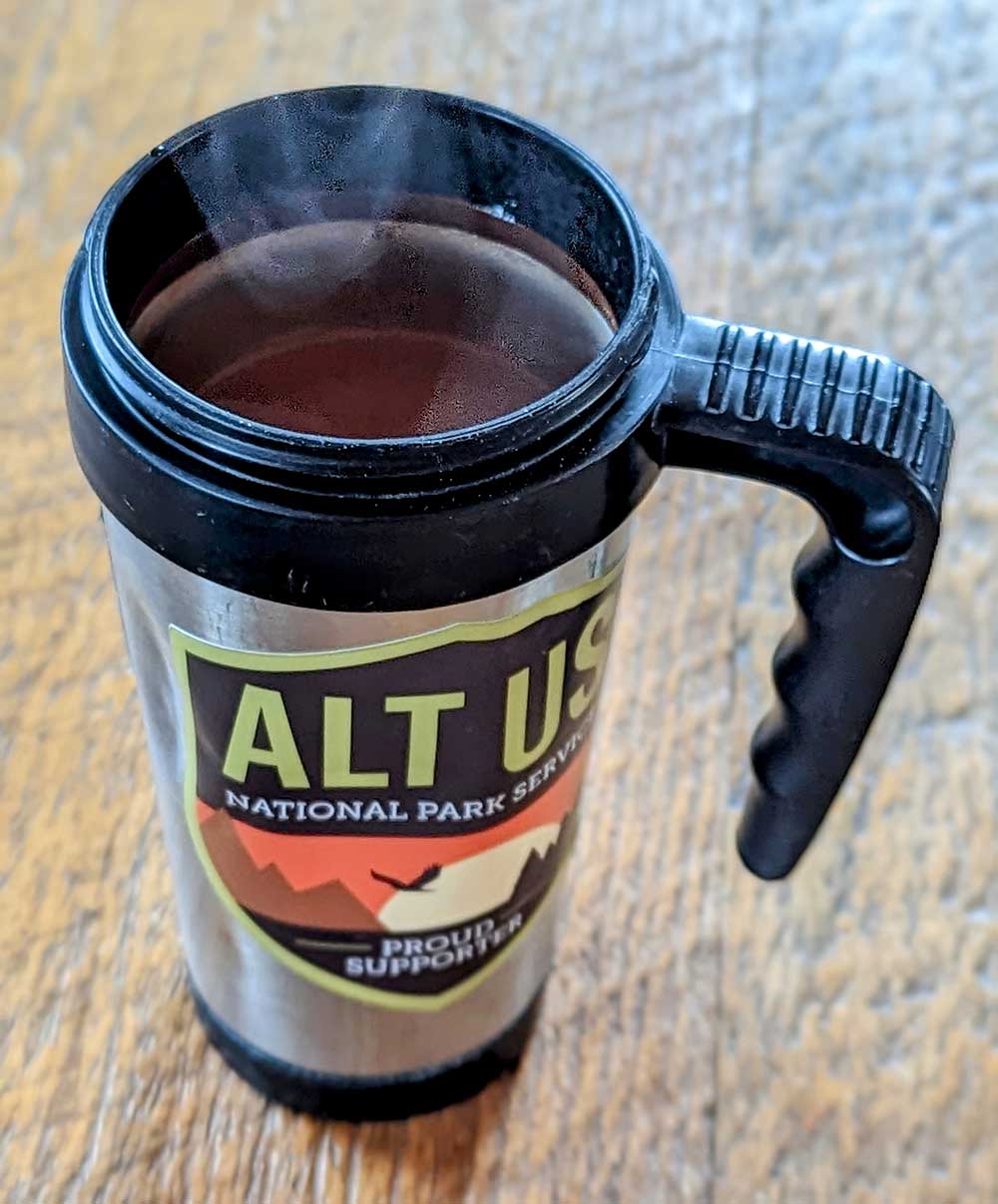 Dented up stainless steel coffee mug full of hot First Ascent Instant Coffee. Steam rises from the cup.