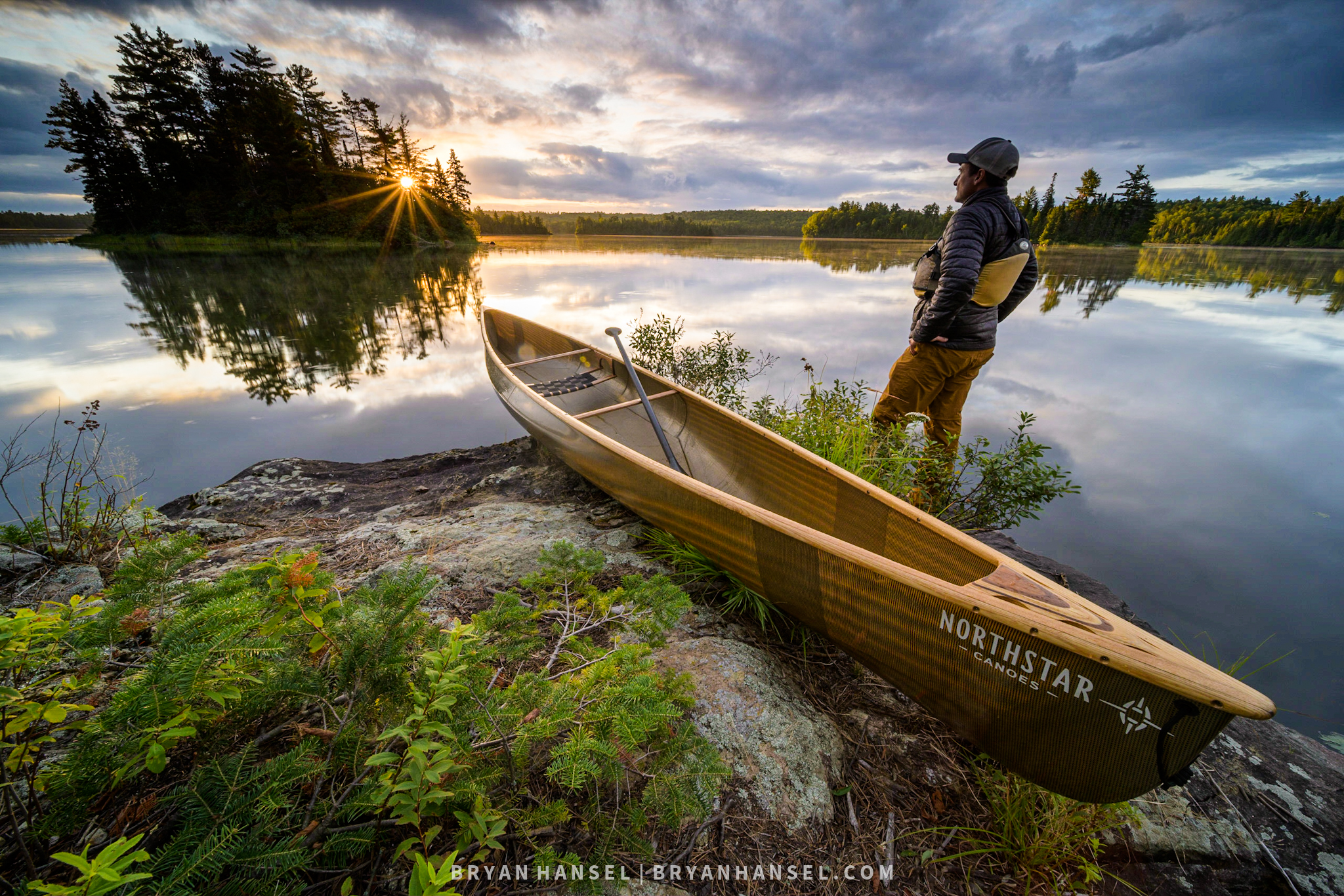 A man standing next to canoe on a Canadian shield Rock Beach, watching sunrise over an island on a calm Lake.