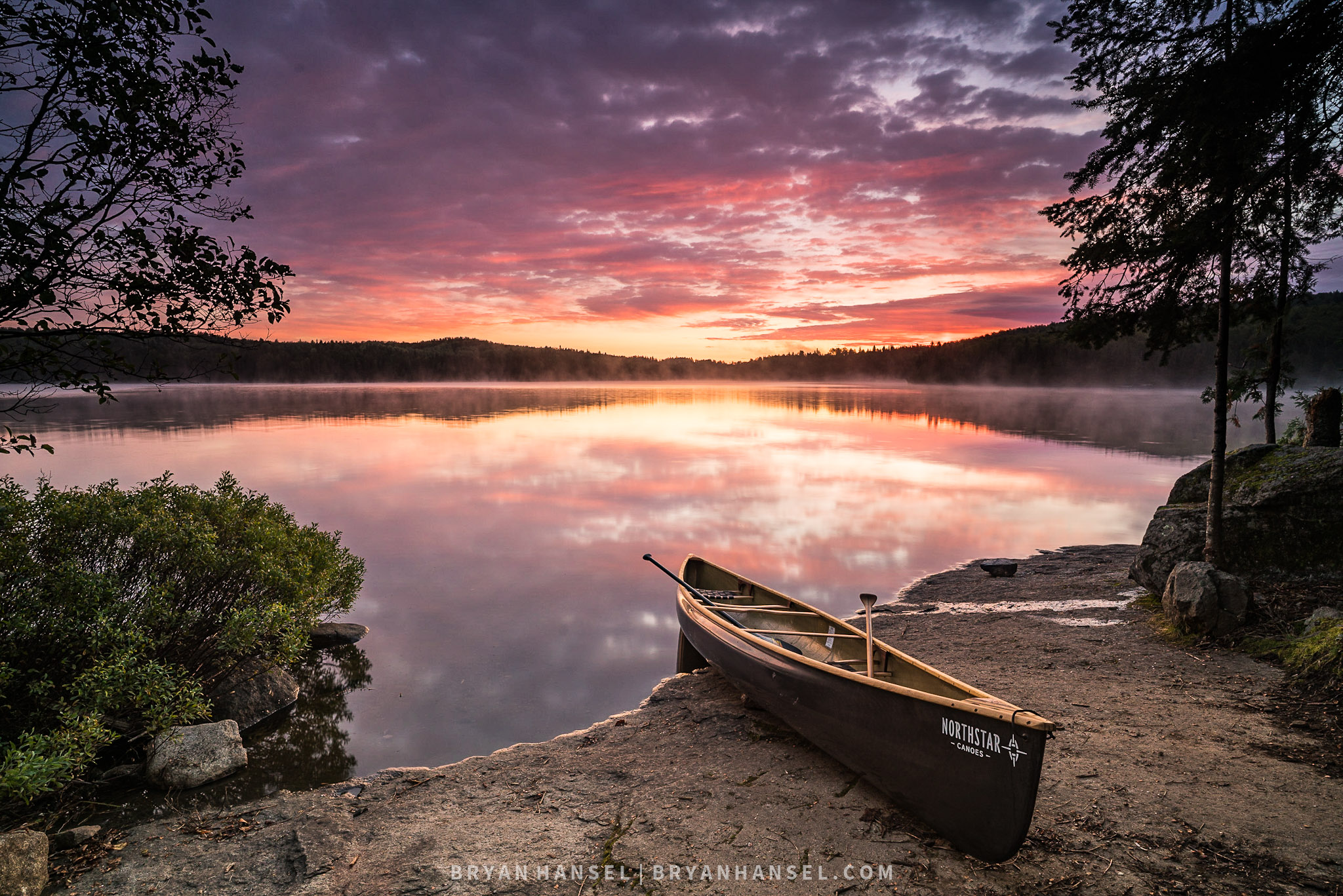 A photo of his new sitting on rock next to a lake at sunrise. The sky is full of red clouds and the reflecting on a perfectly calm water. The picture is framed in by trees on each side. The canoe is black.